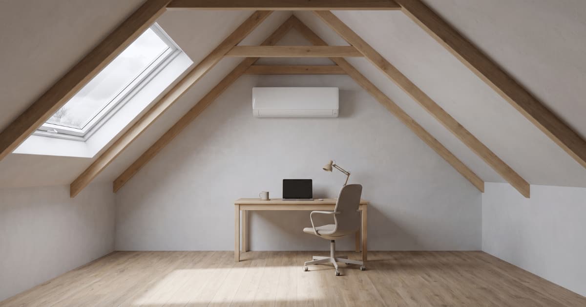 Een witte airco-binnenunit hangt hoog op de kopse wand van een minimalistisch zolderkantoor met zichtbare houten dakbalken, een houten bureau met laptop en bureaulamp en een dakraam dat daglicht binnenlaat.