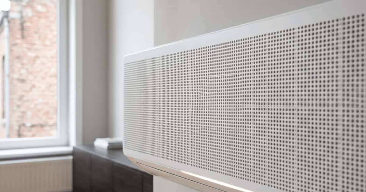 Een witte airco-binnenunit met een fijn geperforeerd frontpaneel hangt aan de muur naast een raam, met een bakstenen gevel zichtbaar op de achtergrond.