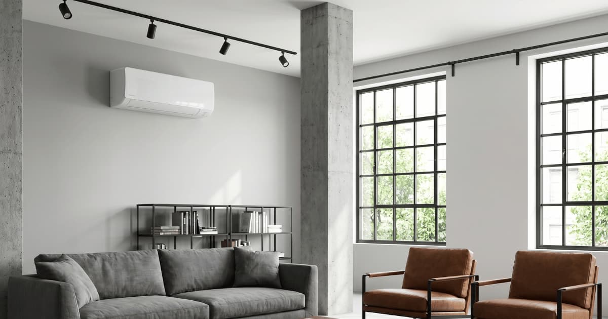 Witte airco-binnenunit hoog gemonteerd op een lichtgrijze muur in een ruime loftwoonkamer met betonnen kolom, zwarte stalen raampartijen en een grijze stoffen bank.
