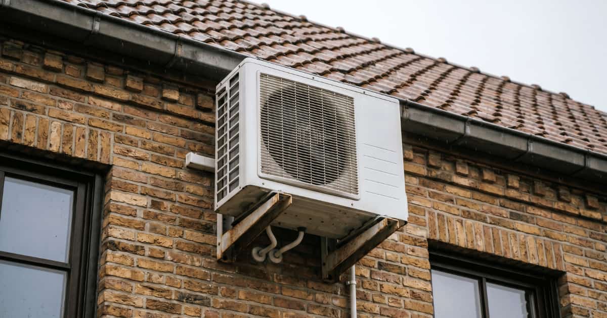 Witte airco buitenunit gemonteerd op een metalen muurbeugel hoog aan een okerkleurige bakstenen gevel net onder de dakgoot met bruine dakpannen en een bewolkte lucht op de achtergrond.