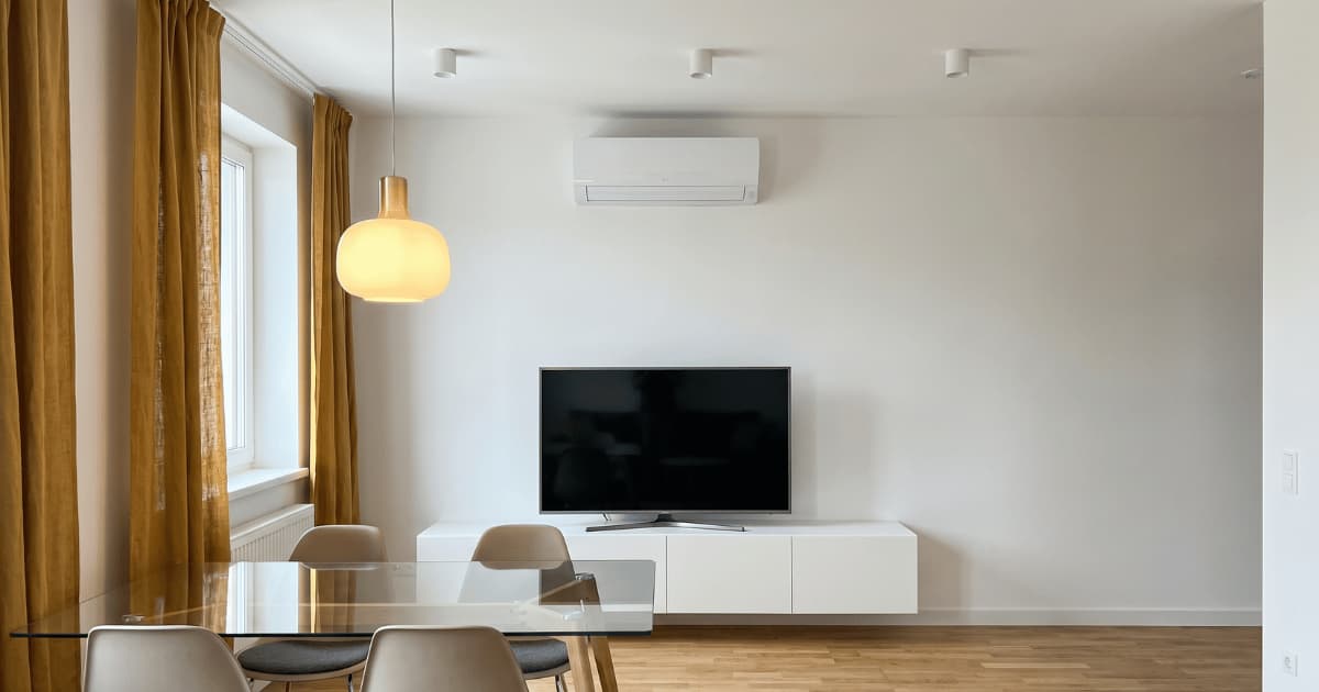 Een witte airco-wandunit hangt hoog boven een televisie op een wit dressoir in een moderne woonkamer met houten vloer, okergele gordijnen en een glazen eettafel.