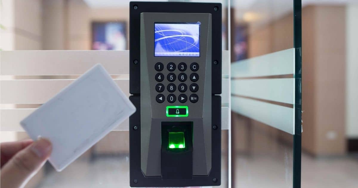 Een hand houdt een witte kaart voor een grijze badgelezer met zwart numeriek toetsenbord. De badgelezer is bevestigd tegen een glazen deur.