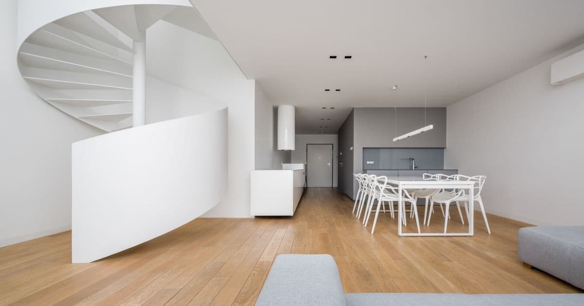 Witte draaitrap trap in een moderne woonkamer. In de woonkamer staat een witte tafel met zes stoelen, een grijze keuken en een grijze zetel.