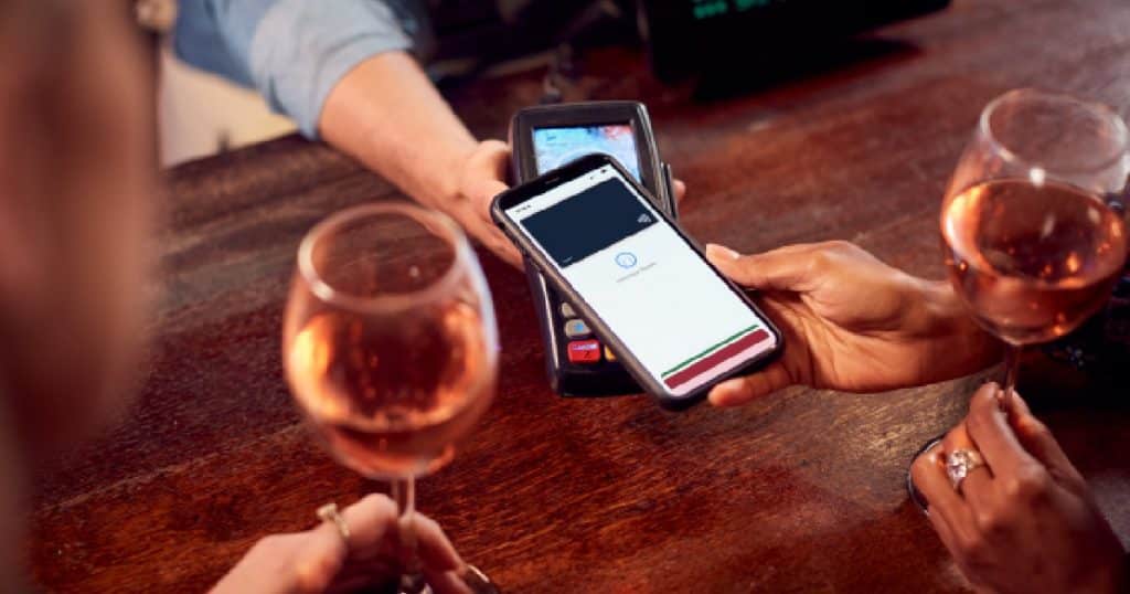 Een dame houdt een wijnglas met rosé vast terwijl ze met haar smartphone afrekent via een mobiele betaalterminal die verbonden is met de witte kassa van een horecazaak.