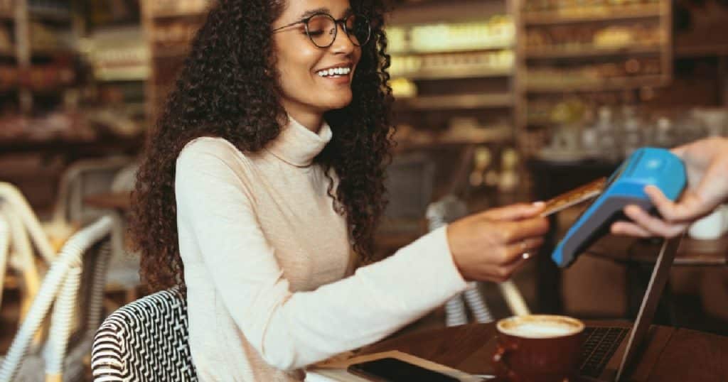 Een dame met bril en een witte trui rekent contactloos af met haar bankkaart via een mobiele betaalterminal die gelinkt is aan de witte kassa van een horecazaak. Voor de dame staan haar laptop en een kop cappuccino.