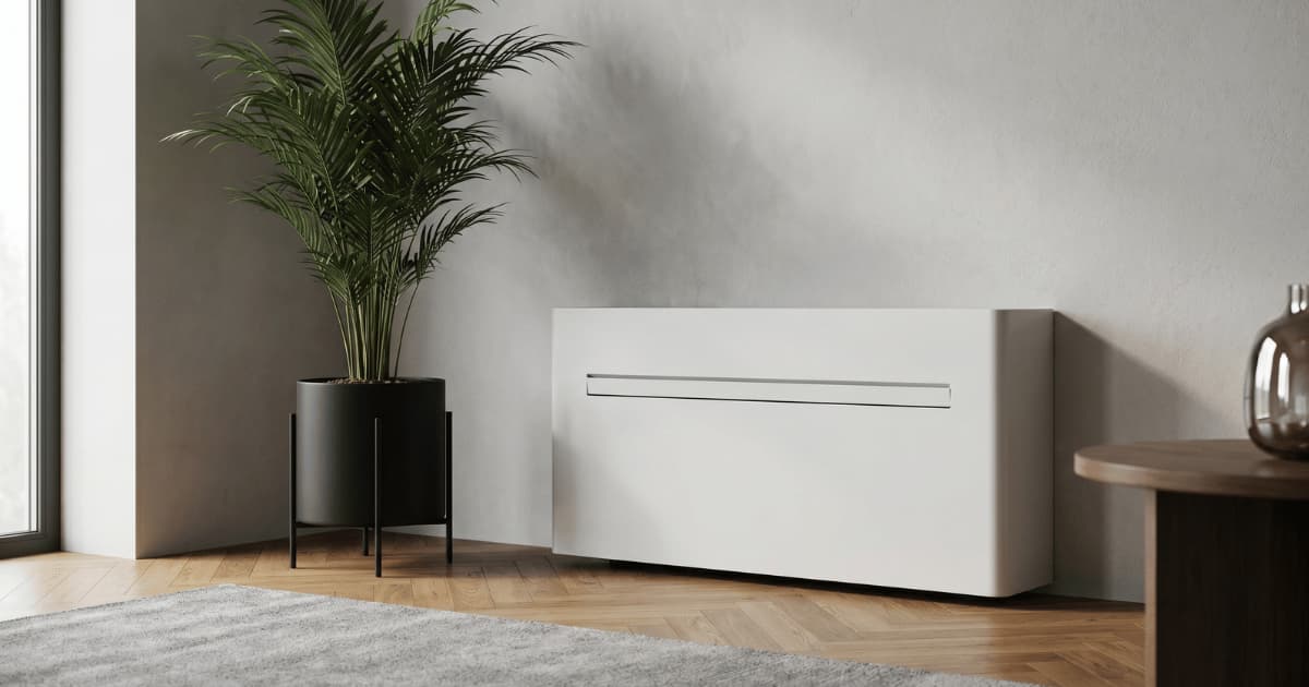 Een witte monoblock airco hangt laag aan een lichtgrijze muur in een woonkamer met visgraat parketvloer en een groene palmplant in een zwarte pot op metalen poten.