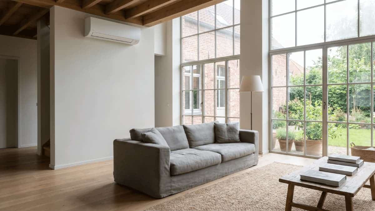 Een witte wandgemonteerde split-airco hangt hoog op de muur van een ruime woonkamer met zichtbare houten dakbalken, een grijze stoffen zitbank en grote ramen die uitkijken op een bakstenen gevel en groene tuin.