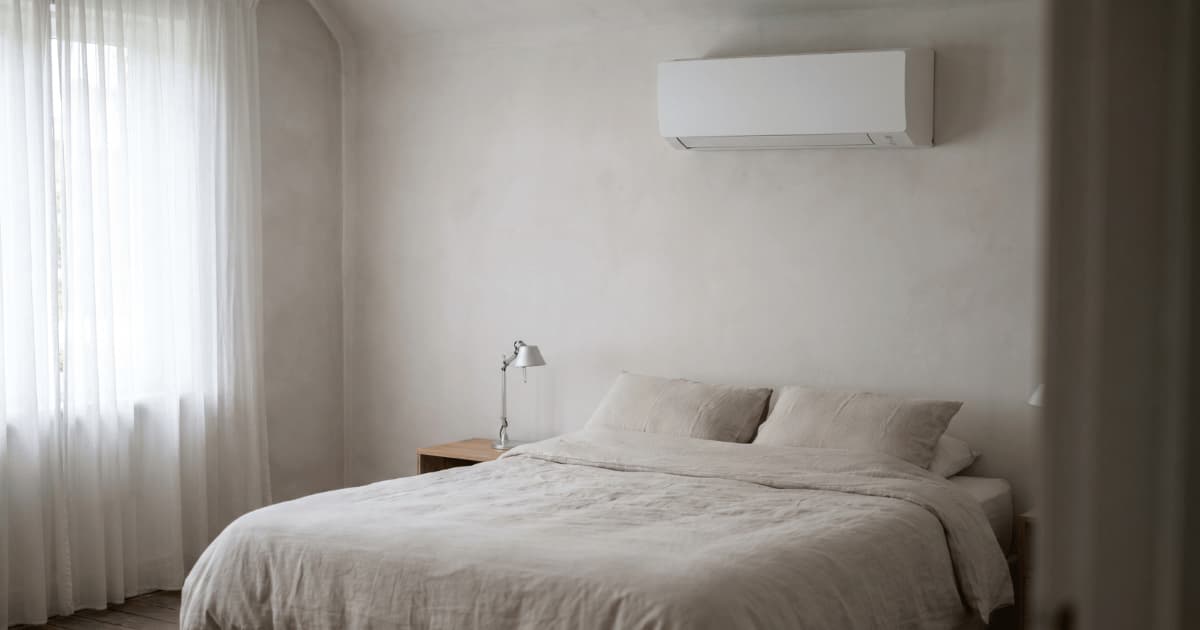 Een witte wandgemonteerde airco-unit hangt hoog aan een lichtgrijze muur boven een tweepersoonsbed met beige linnen beddengoed in een minimalistische slaapkamer met houten nachtkastje, metalen leeslamp en doorschijnende witte gordijnen.