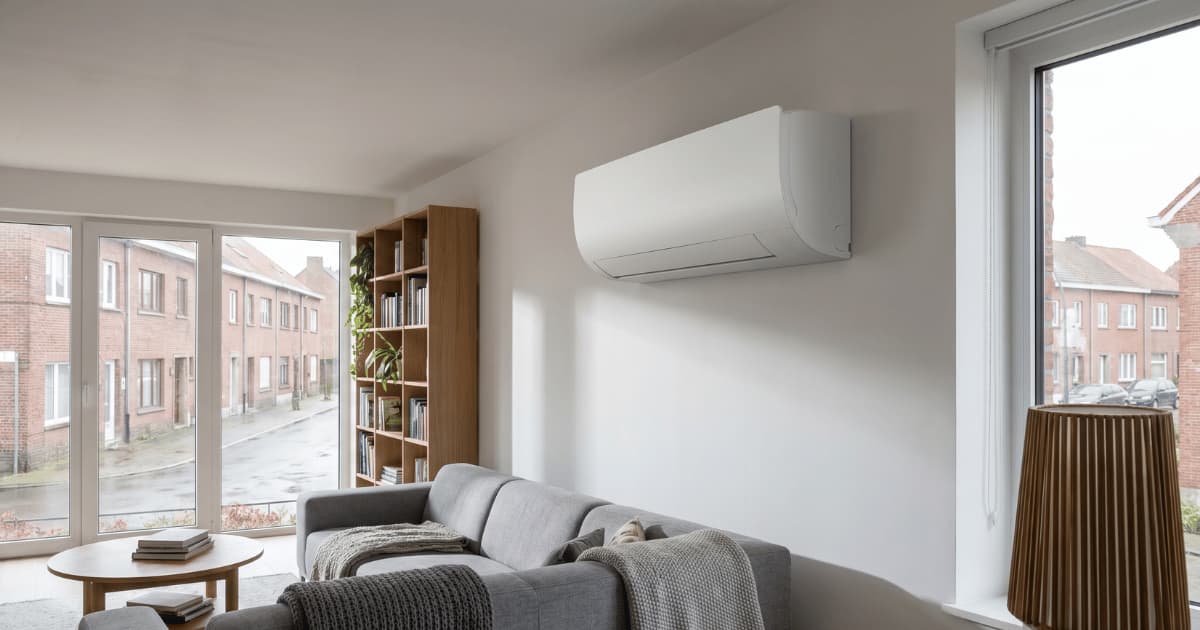 Een witte wandairco hangt aan de muur van een moderne woonkamer met grijze hoekzetel en houten boekenkast, terwijl door de grote ramen en glazen schuifdeur een straat met Vlaamse bakstenen rijwoningen zichtbaar is op een bewolkte dag.