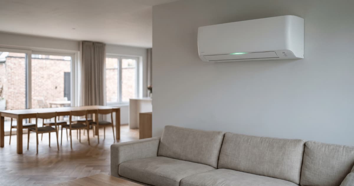 Een witte wandairco met een klein groen indicatielampje hangt boven een beige zitbank in een ruime open woonkamer met houten eettafel en stoelen op de achtergrond en natuurlijk daglicht door grote ramen met zicht op een bakstenen gevel.
