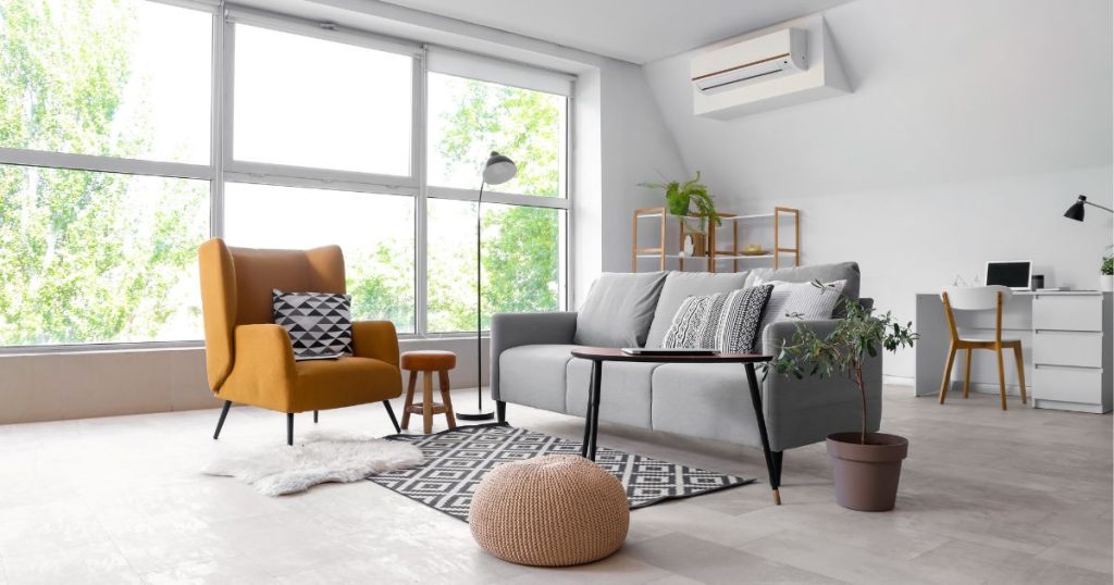 Een woonkamer met linksboven een Mitsubishi Electric airco, een grijze vloer, grijze zetels, een houten bijzettafeltje en een zwarte designlamp. 