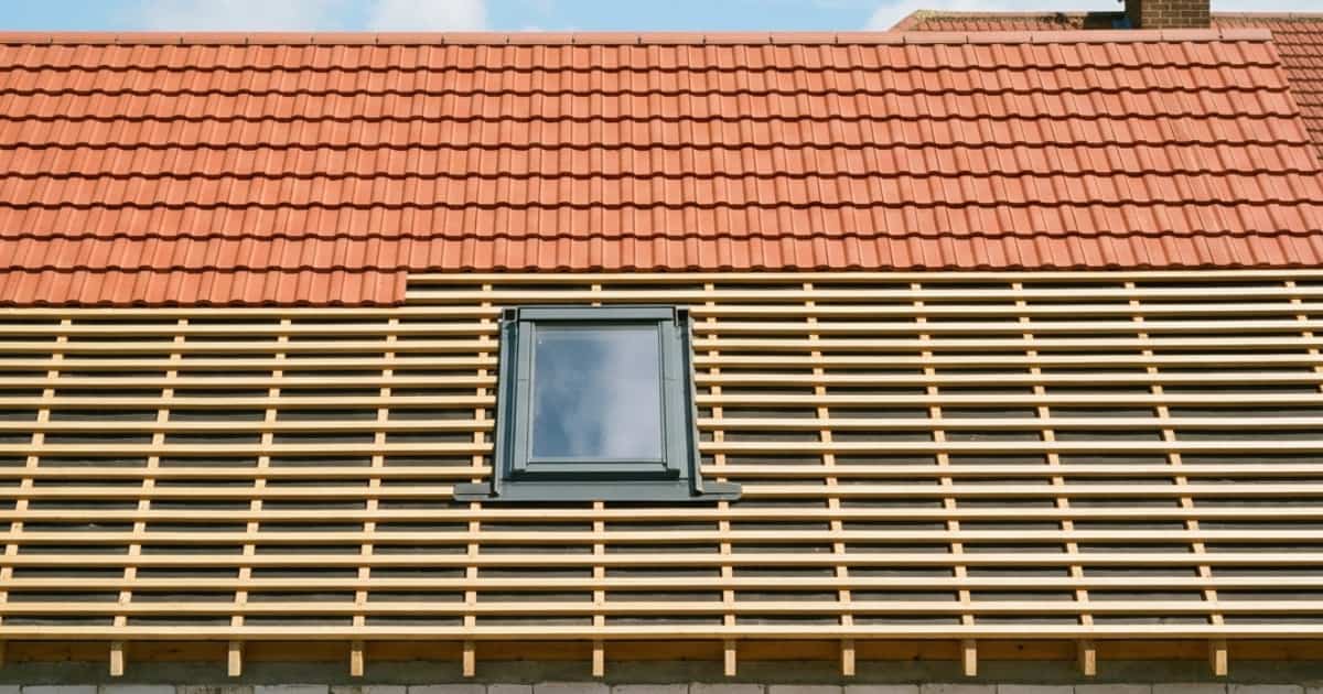 Een zadeldak in aanbouw toont de bovenste helft al bedekt met rode keramische dakpannen terwijl de onderste helft nog de kale houten panlatten laat zien met een ingebouwd dakraam met donkergrijs frame in het midden.