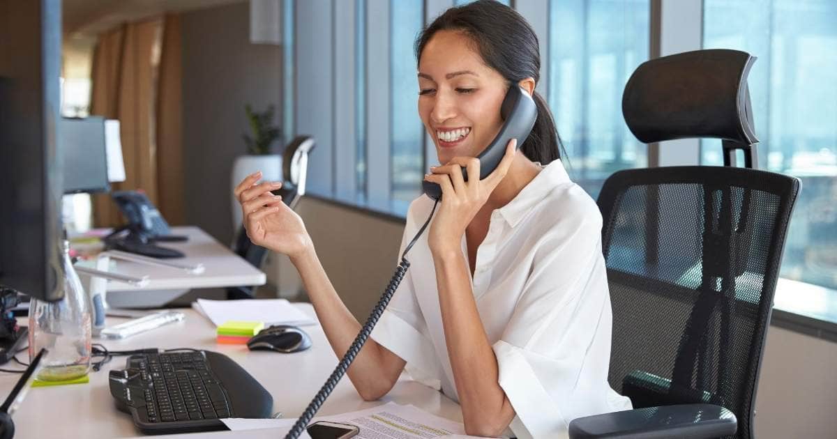 Een dame met lang donkerbruin haar en een wit hemd telefoneert met een zwart telefoontoestel. Ze zit op een zwarte bureaustoel met mesh rug. Op haar witte bureautafel liggen een toetsenbord, computermuis, papieren en post-its.