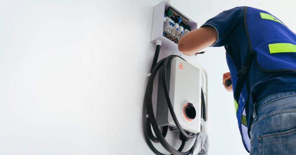 Een installateur is zelf een laadpaal aan het plaatsen bij een klant zelf. Het gaat om een wit wallbox model dat tegen een witte buitengevel bevestigd wordt.