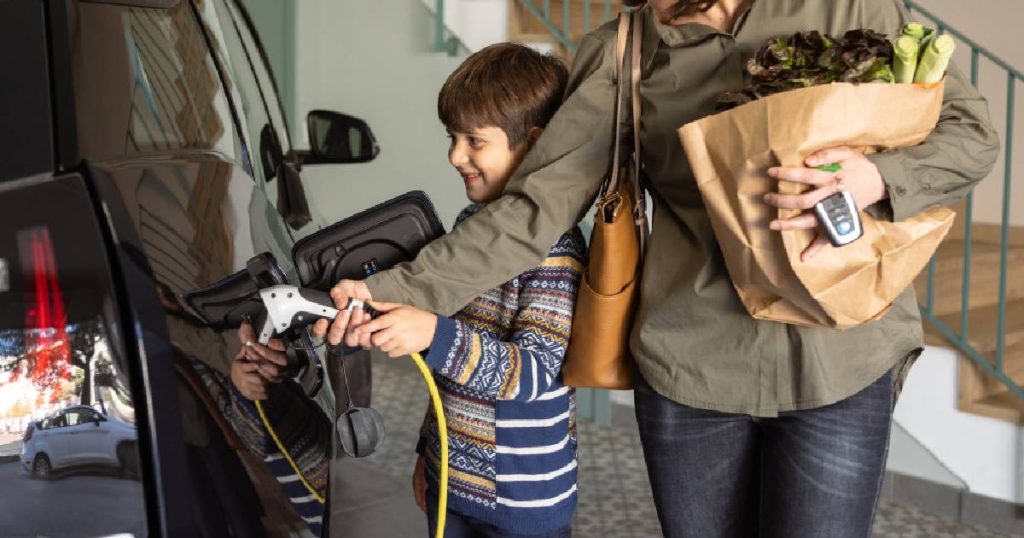 Een moeder en kind pluggen de laadkabel van een laadpaal in een elektrische auto die ze zelf plaatsten. De moeder draagt een papieren tas met boodschappen in haar ene arm en heeft de autosleutels tussen haar vingers. Ze draagt een groene jas en donkere jeansbroek. Het kind draat een gestreepte trui.