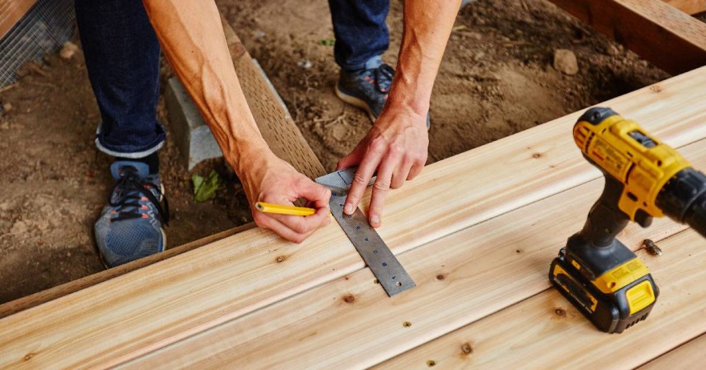 Een man is zelf een veranda aan het maken en verzaagt de houten planken op maat. Op de planken staat een boormachine.