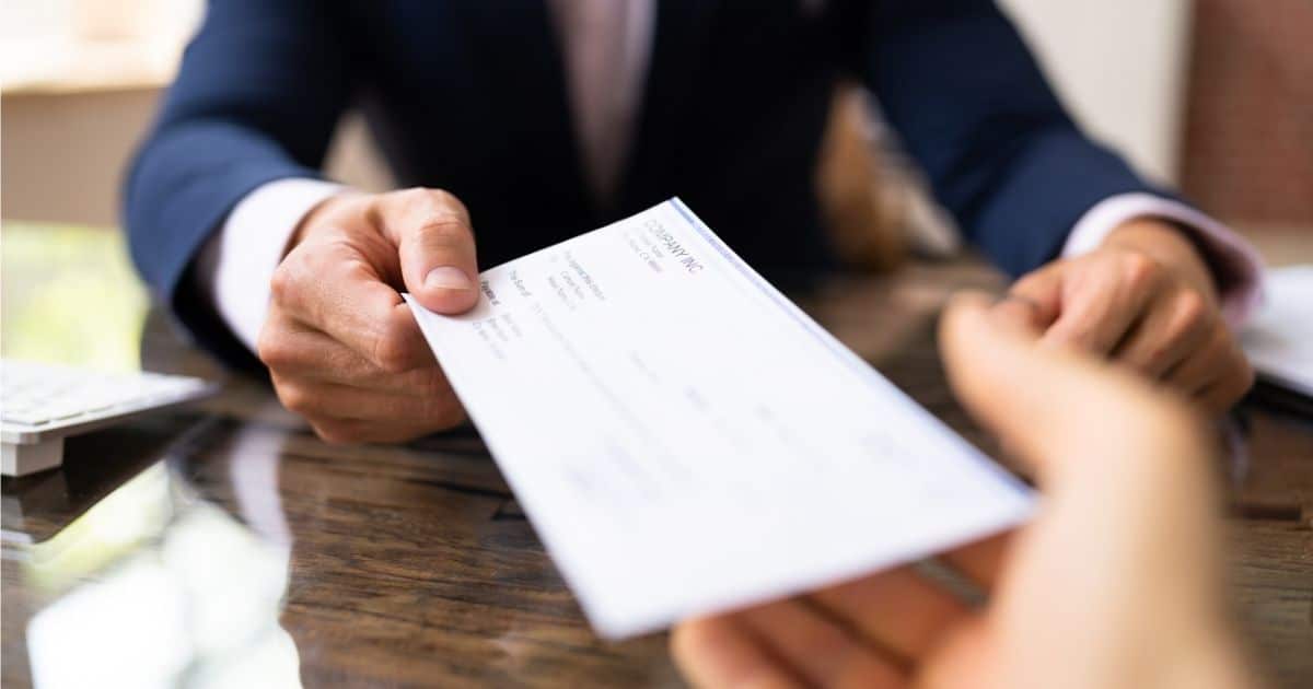 Een man in blauw maatpak met wit hemd en roze das overhandigt een loonfiche. Enkel de schouders, borst en armen van de man zijn zichtbaar. Van de ontvanger van het loonfiche is enkel de rechterhand in beeld.