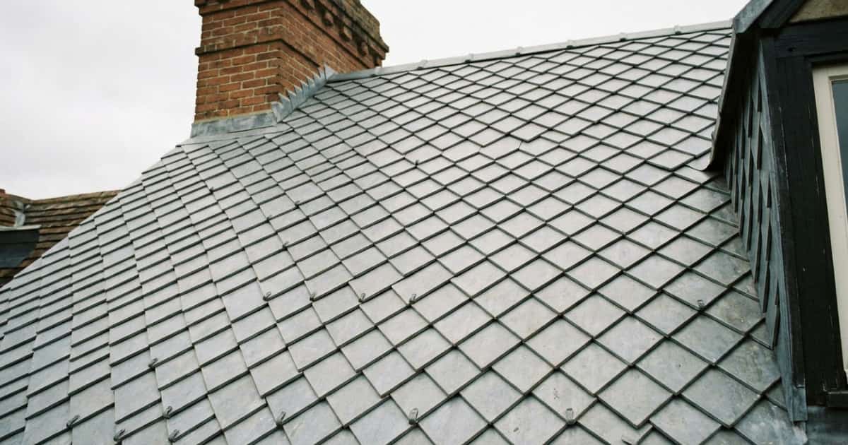Hellend dak bedekt met ruitvormige zinken losanges in een overlappend mozaïekpatroon met zichtbare bevestigingsclips, een bakstenen schoorsteen met zinken aansluitingen nabij de nok en een dakkapel met donker houten kozijn rechts in beeld.