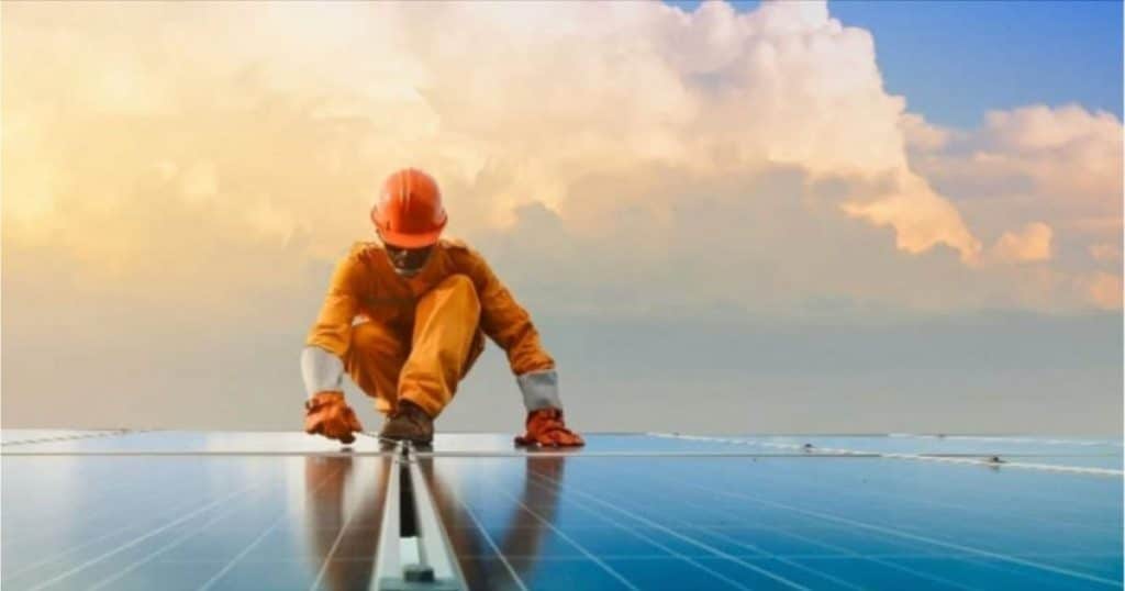 Een man die zonnepanelen in West-Vlaanderen plaatst op een plat dak. De man draagt oranje werkkledij en een helm.