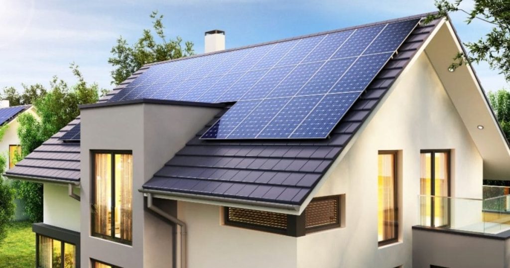 De bovenkant van een huis met een dak vol zonnepanelen. Het is zonnig buiten en het hele huis is verlicht. De muren van het huis zijn wit en het dak onder de zonnepanelen is donkergrijs