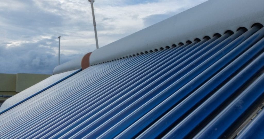 Een foto van een zonneboiler op een dak van dichtbij. De zonneboiler is blauw en de bovenkant wit. De lucht erboven is erg bewolkt en grijs