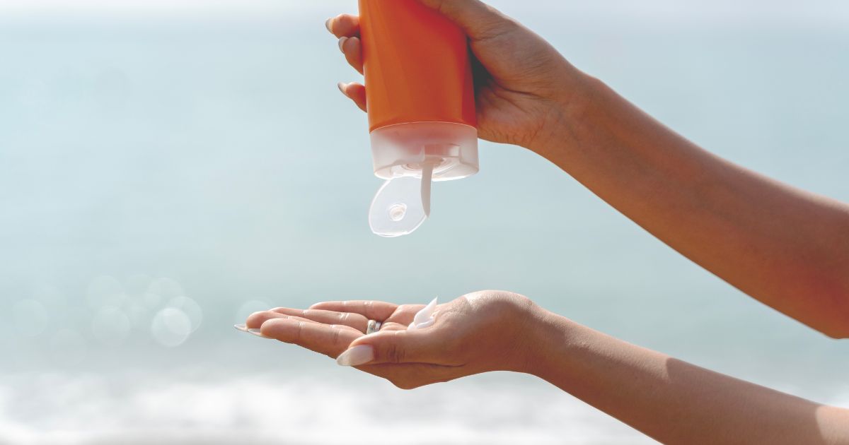 Een hand knijpt in een oranje flacon zonnecrème: eronder een open hand om de zonnecrème op te vangen. De zonnige zee op de achtergrond.