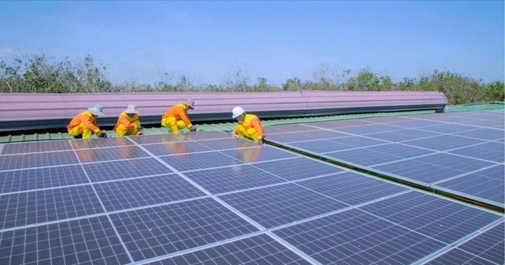 Vier vakmannen zijn gekleed in gele werkkledij. Ze zijn zonnepanelen aan het installeren op het dak van een groot bedrijfspand.