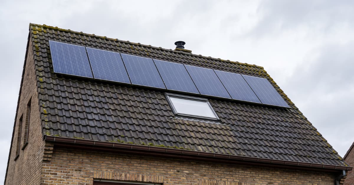Rij donkerblauwe zonnepanelen gemonteerd op het bovenste deel van een hellend dak met verweerde antracietkleurige dakpannen en mosgroei boven een dakraam op een Vlaamse bakstenen gevel onder een bewolkte hemel.