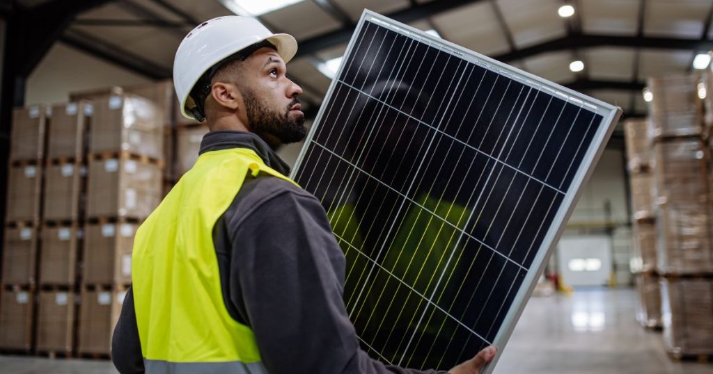 Een man met witte veiligheidshelm en geel veiligheidsvestje draagt een oud zonnepaneel weg van een bestaande zonnepaneelinstallatie die vervangen moet worden.