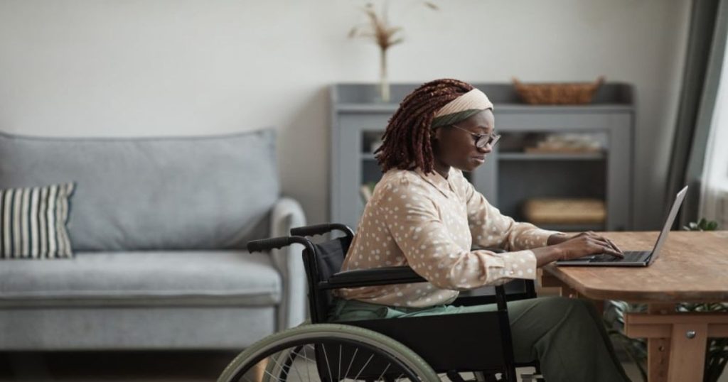 Een dame in een rolstoel typt op een laptop aan een tafel. Op de achtergrond staan een grijze sofa en een vitrinekast.