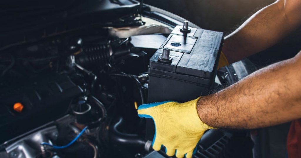 Een automonteur met geel met blauwe werkhandschoenen haalt een oude loodzuurbatterij uit een auto.