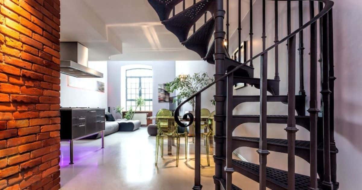 Zijaanzicht van een donkere ruimtebesparende wenteltrap. Deze staat tegen een paarse muur. In de kamer is een kleine keuken, een woonkamer met zetels en een eettafel.