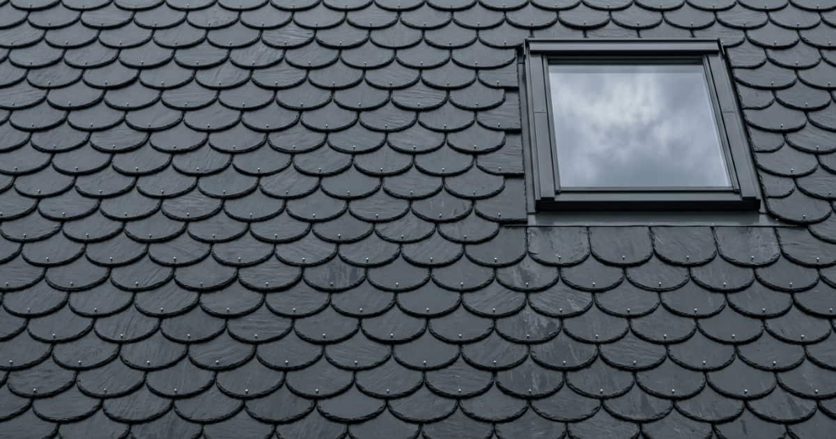 Zwarte natuurleien met een afgeronde onderkant liggen in een verspringend schubbenpatroon rond een dakraam dat de bewolkte lucht weerspiegelt.
