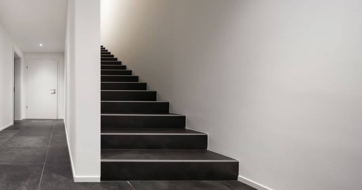 Een trap met zwarte overzettreden in betonlook in een wit geschilderde hal. De treden zijn voorzien van antislip strips, en de trap geeft uit op een stijlvolle vloer met zwarte, natuurstenen tegels.