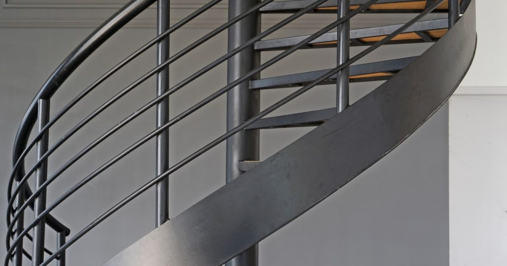 Een wenteltrap in metaal met een moderne twist. De grijze kleur van het frame van de trap sluit mooi aan op de grijs geverfde muren.