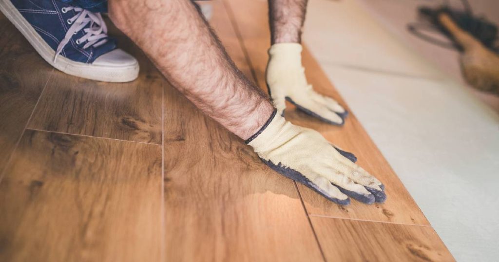 Een man met blauwe sneakers en geel met grijze handschoenen legt zwevend honignbruin parket op een grijze ondervloer.